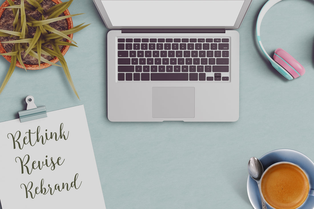 A piece of paper on a clipboard says 'Rethink, Revise, Rebrand'. It sits to the left of an open laptop and a cup of coffee.
